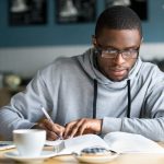 Focused millennial african american student in glasses making notes writing down information from book in cafe preparing for test or exam, young serious black man studying or working in coffee house