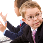 child in glasses and a suit with tie sits next to another child in business apparel, both clearly directing an office