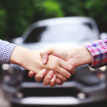 Two people shake hands with a car in the background